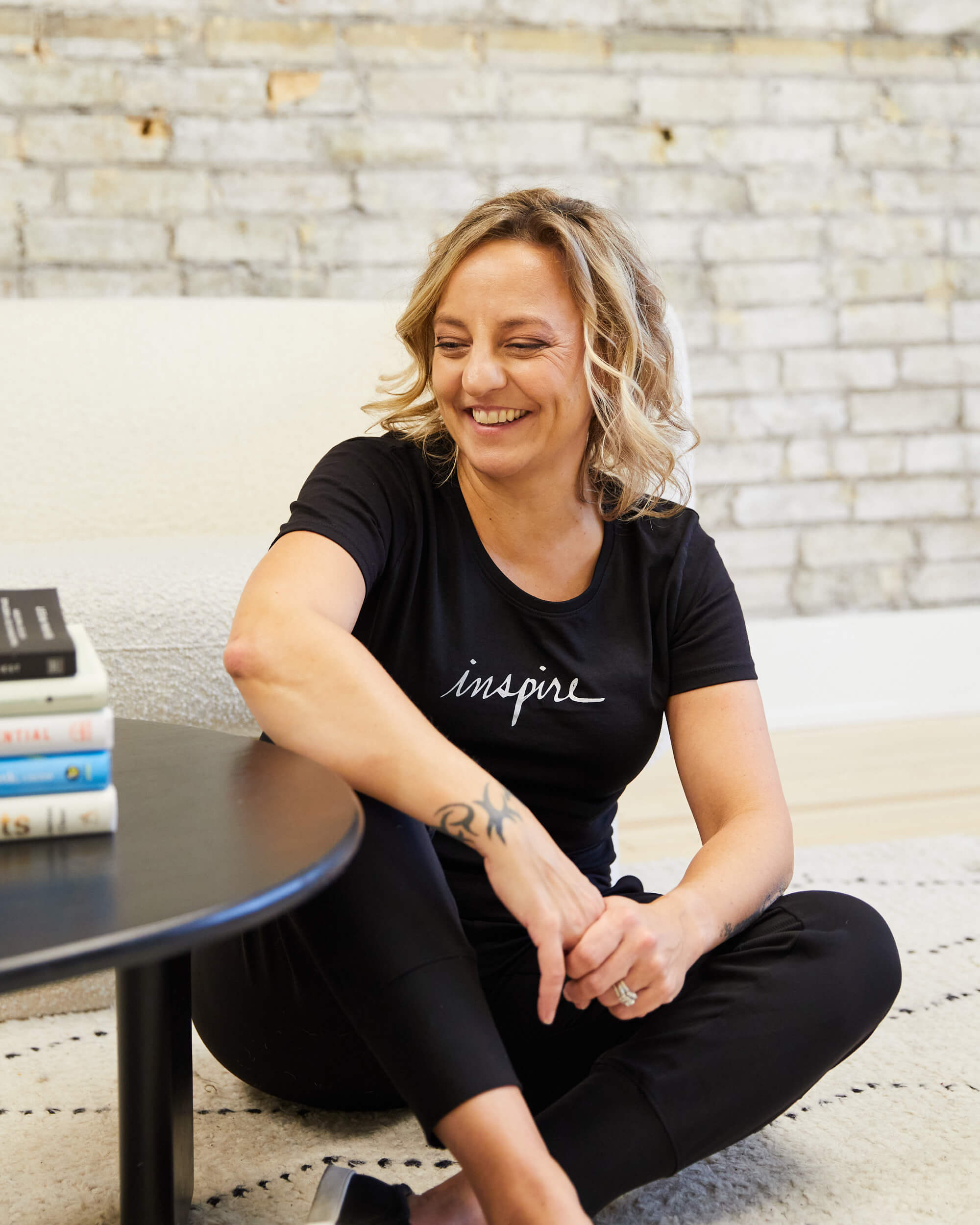 Michelle wears a black t-shirt with the word "inspire" on it. She is sitting on the floor, cross-legged and leaning on a coffee table while laughing.