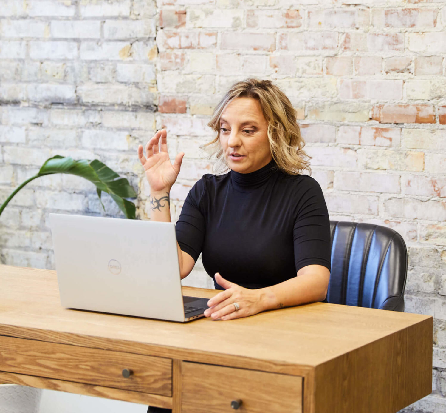 Michelle sits behind a wooden desk and has her laptop open. She is facing the computer and speaking to someone virtually.