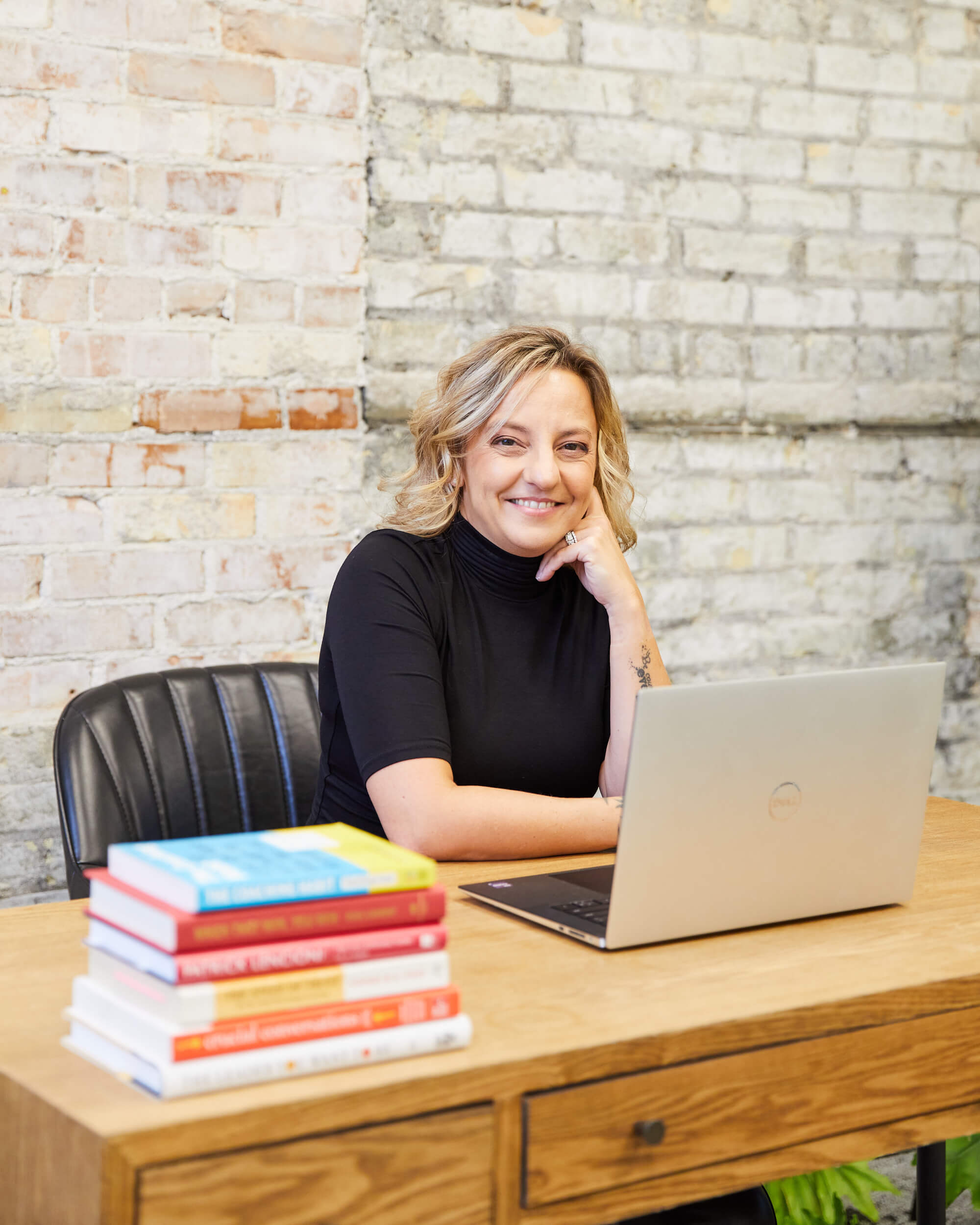 Michelle sits behind a wooden desk with her laptop open. She is looking across to the camera and smiling with a stack of leadership books beside her.