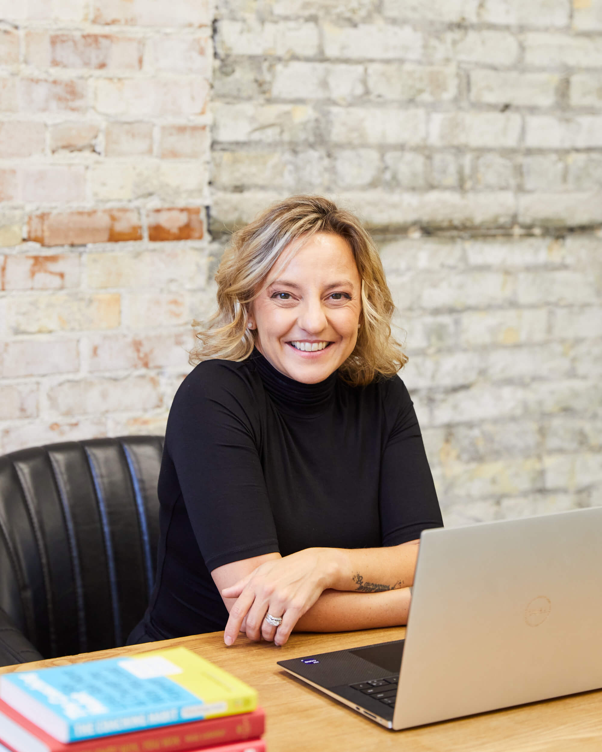 Michelle Martin is sitting behind a wooden desk with her laptop open. She has a stack of books beside her and is looking at the camera and smiling.